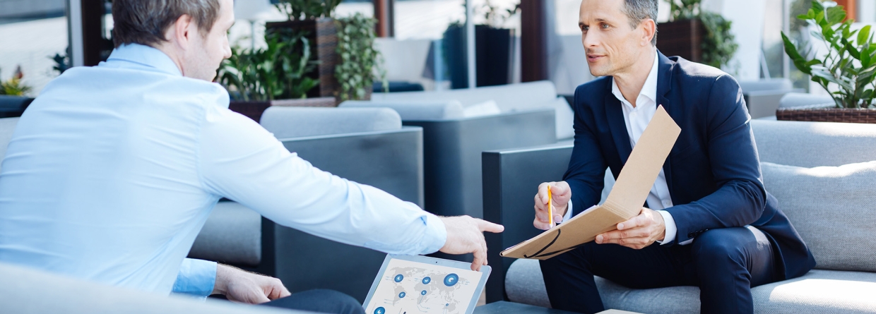 Man in pak wijst naar document in zijn hand aan man tegenover hem met een laptop voor zich